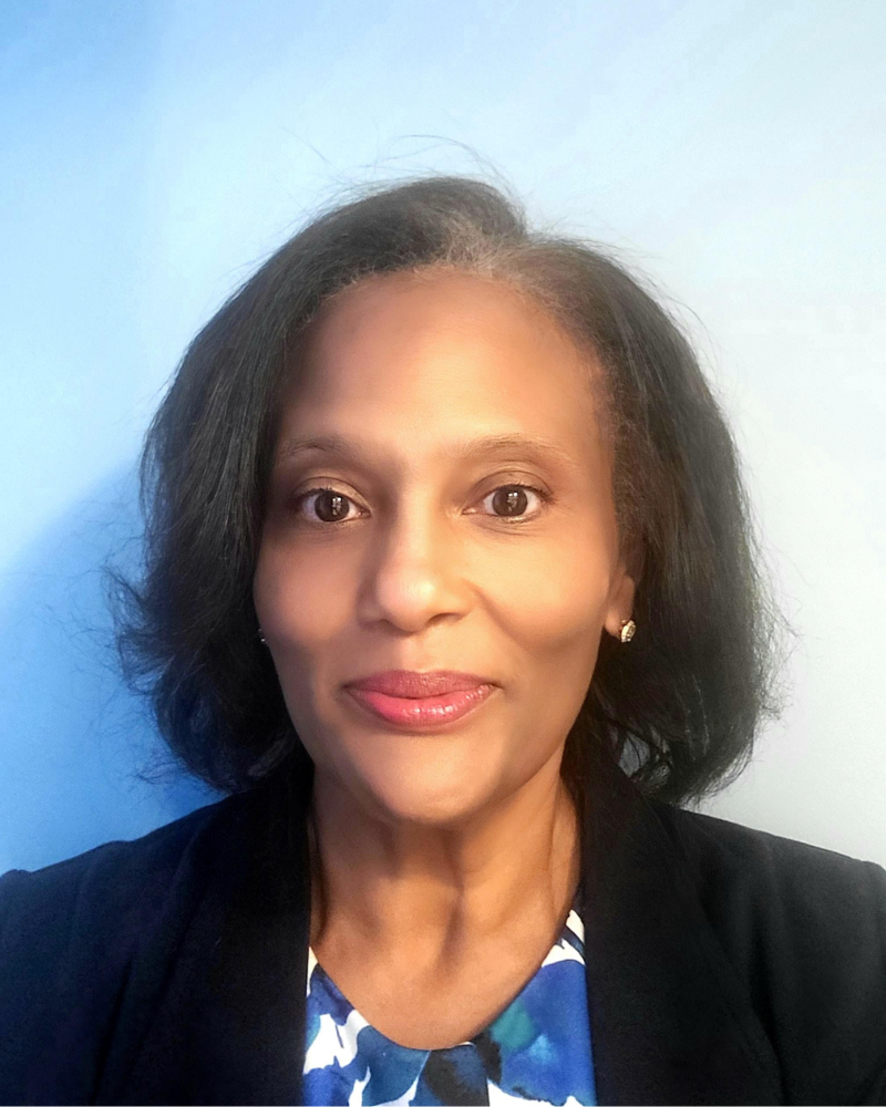 **Alt text:** Portrait of a woman with shoulder-length dark hair wearing a dark blazer and patterned top, photographed against a soft blue background.